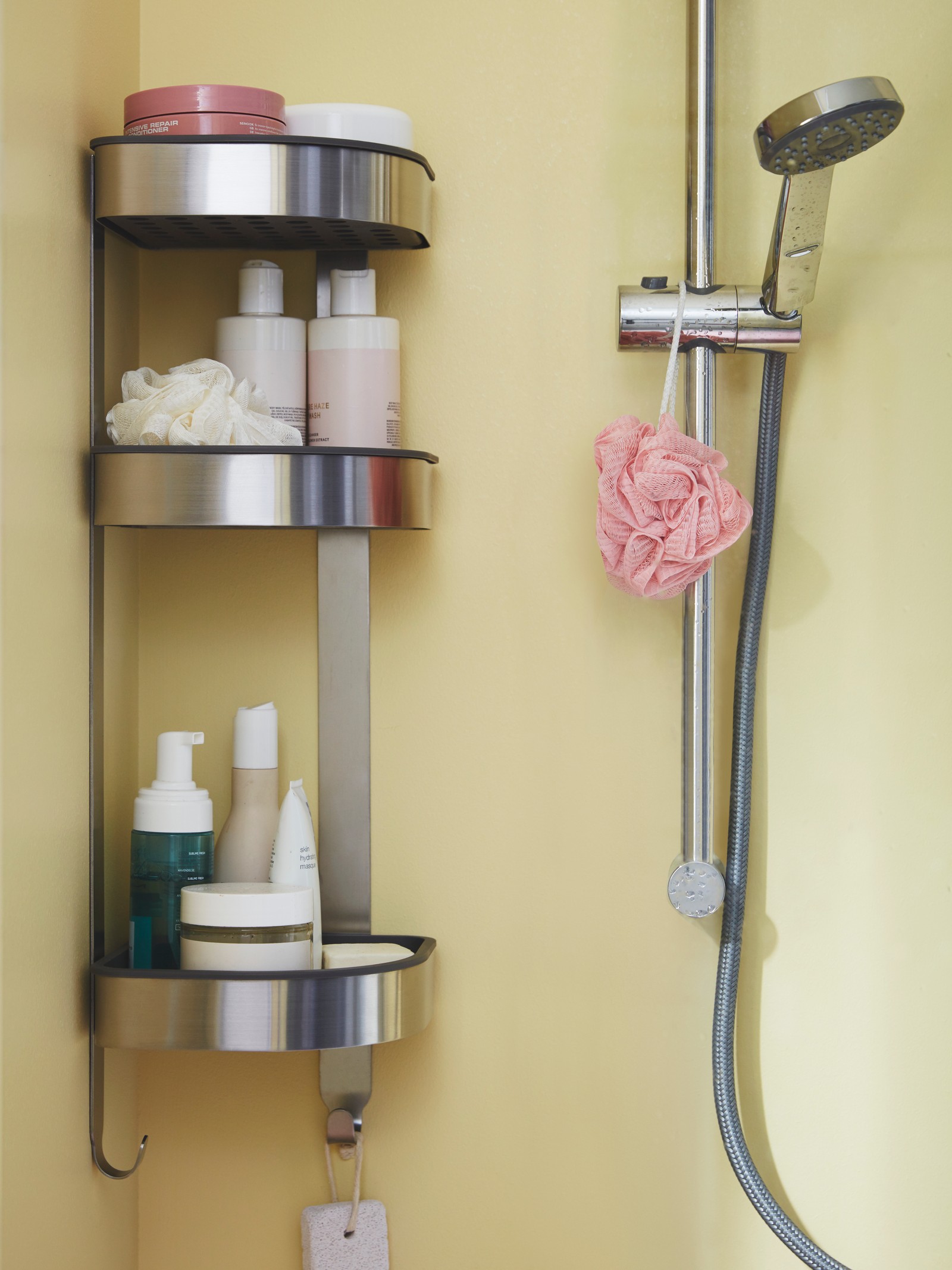 Un organizador vertical cromada VALLAMOSSE plateada con artículos de ducha en sus repisas y al costado una ducha IKEA.