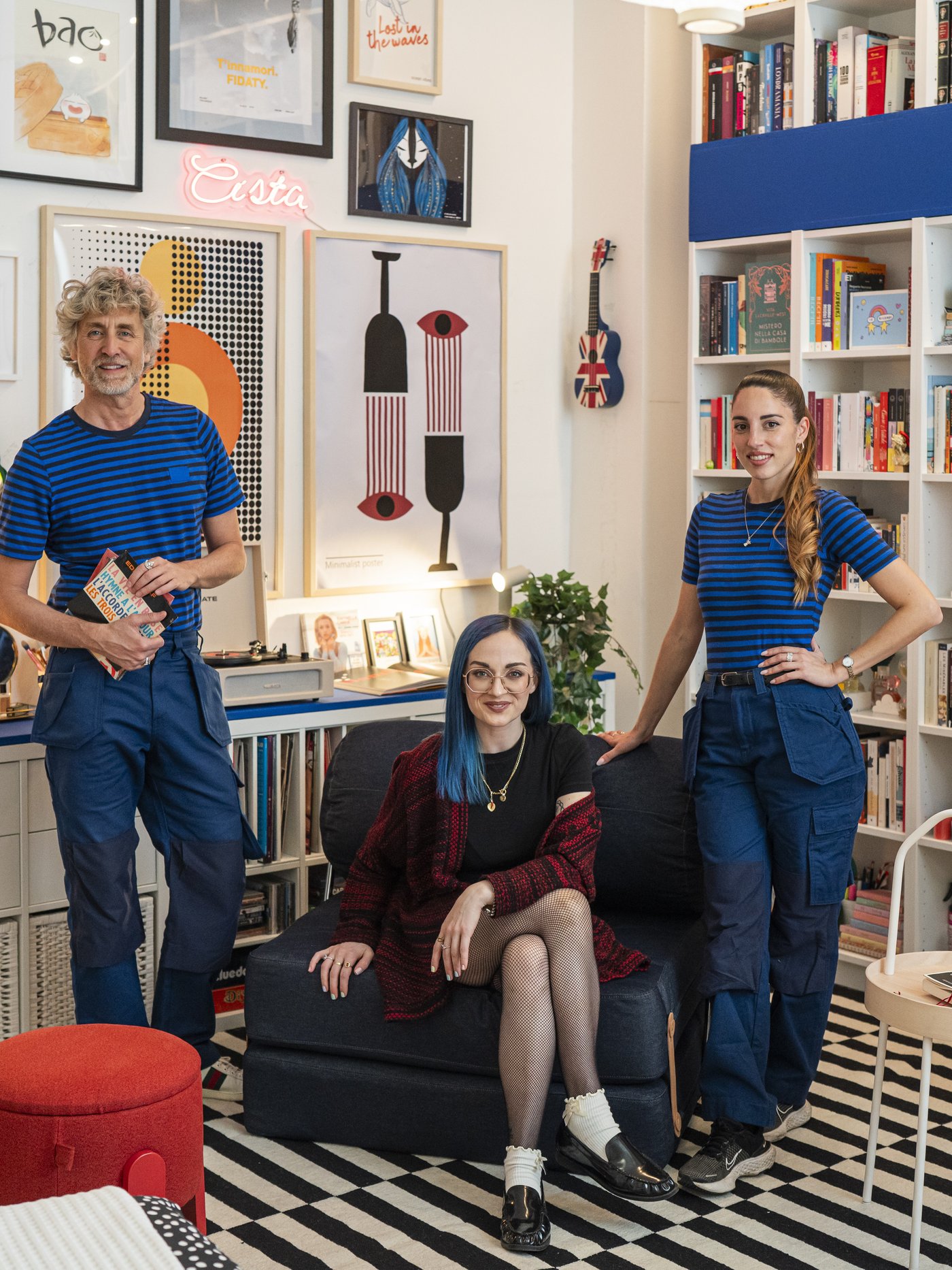 Due interior designer IKEA in divisa blu posano in un soggiorno colorato e pieno di libri accanto a Teresa Agostini su una poltrona scura.