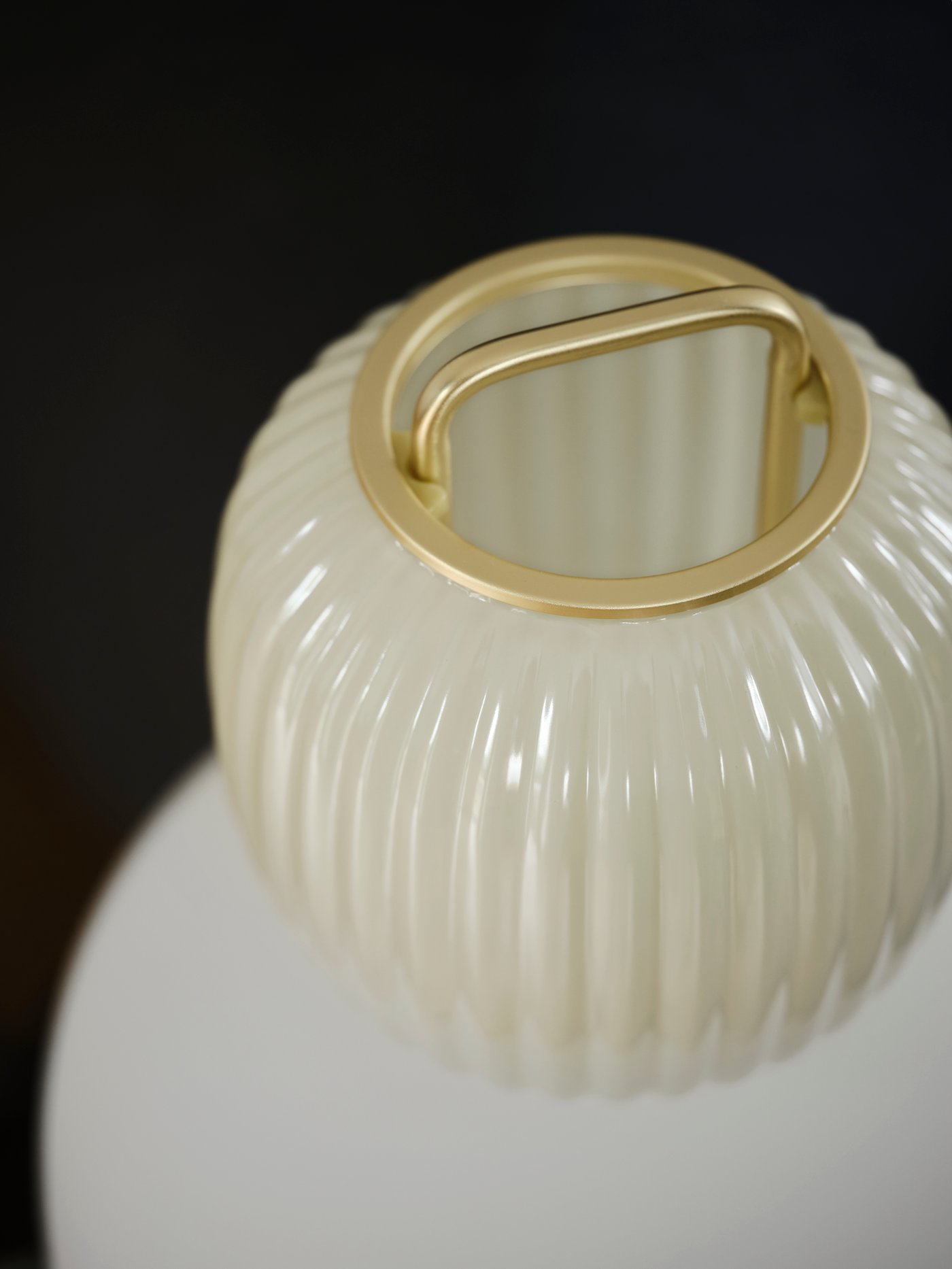 The top part of a white/light green glass PILBLIXT lamp with gold effect metal in front of a black wall.
