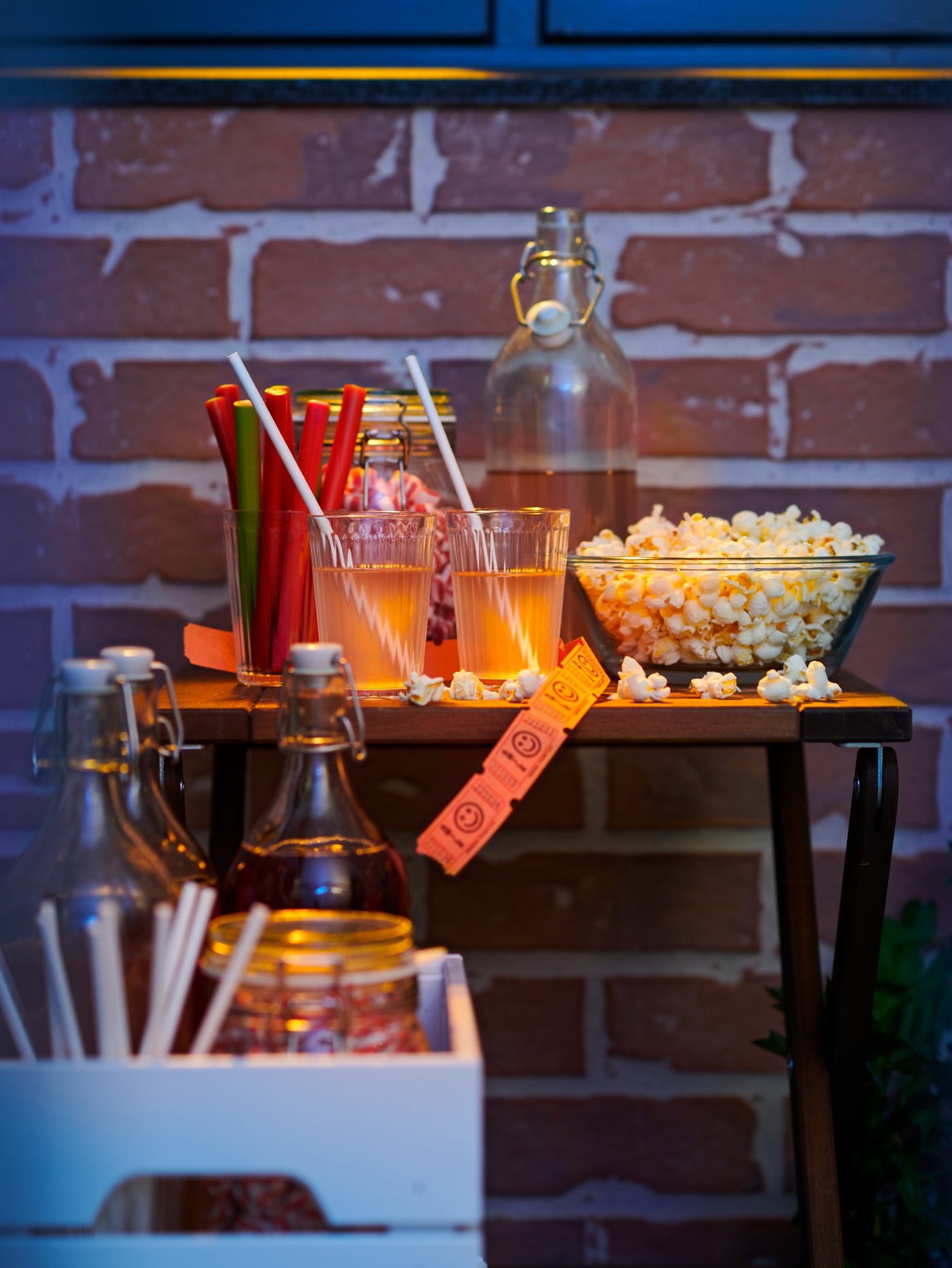 Una mesa con cosas para picar en un balcón de ladrillo para disfrutar de una sesión de cine. Hay un cuenco con palomitas y bebidas con pajitas de papel FÖRNYANDE.
