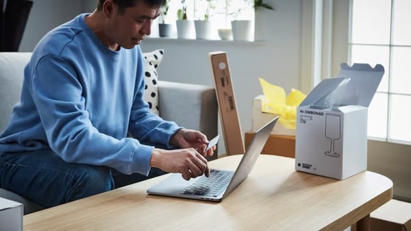 Un uomo con una felpa azzurra è seduto su un divano bianco e utilizza un computer portatile intento a eseguire un pagamento online.