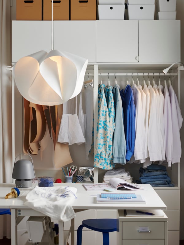 A desk in the foreground and an open PLATSA wardrobe combination in the back, with hanging clothes and sowing projects.