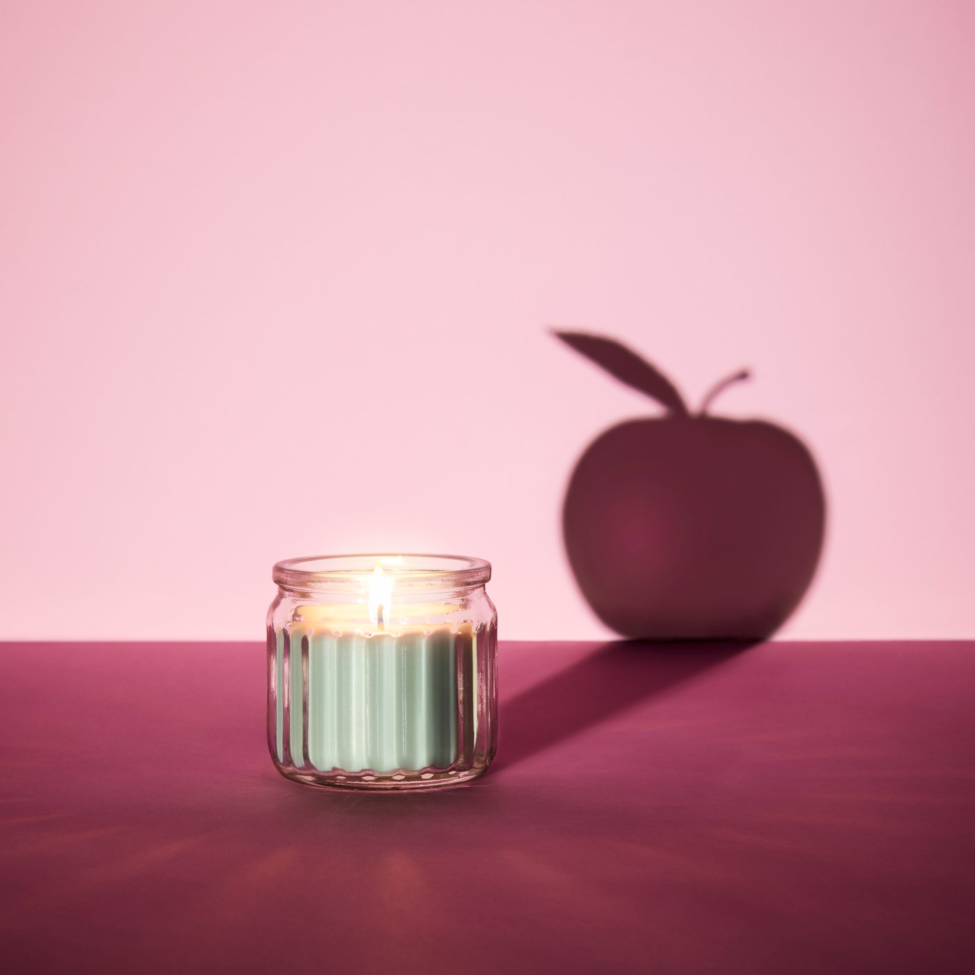 A lit HÖKGÖK scented candle, casting a long, graphic shadow in the shape of an apple.