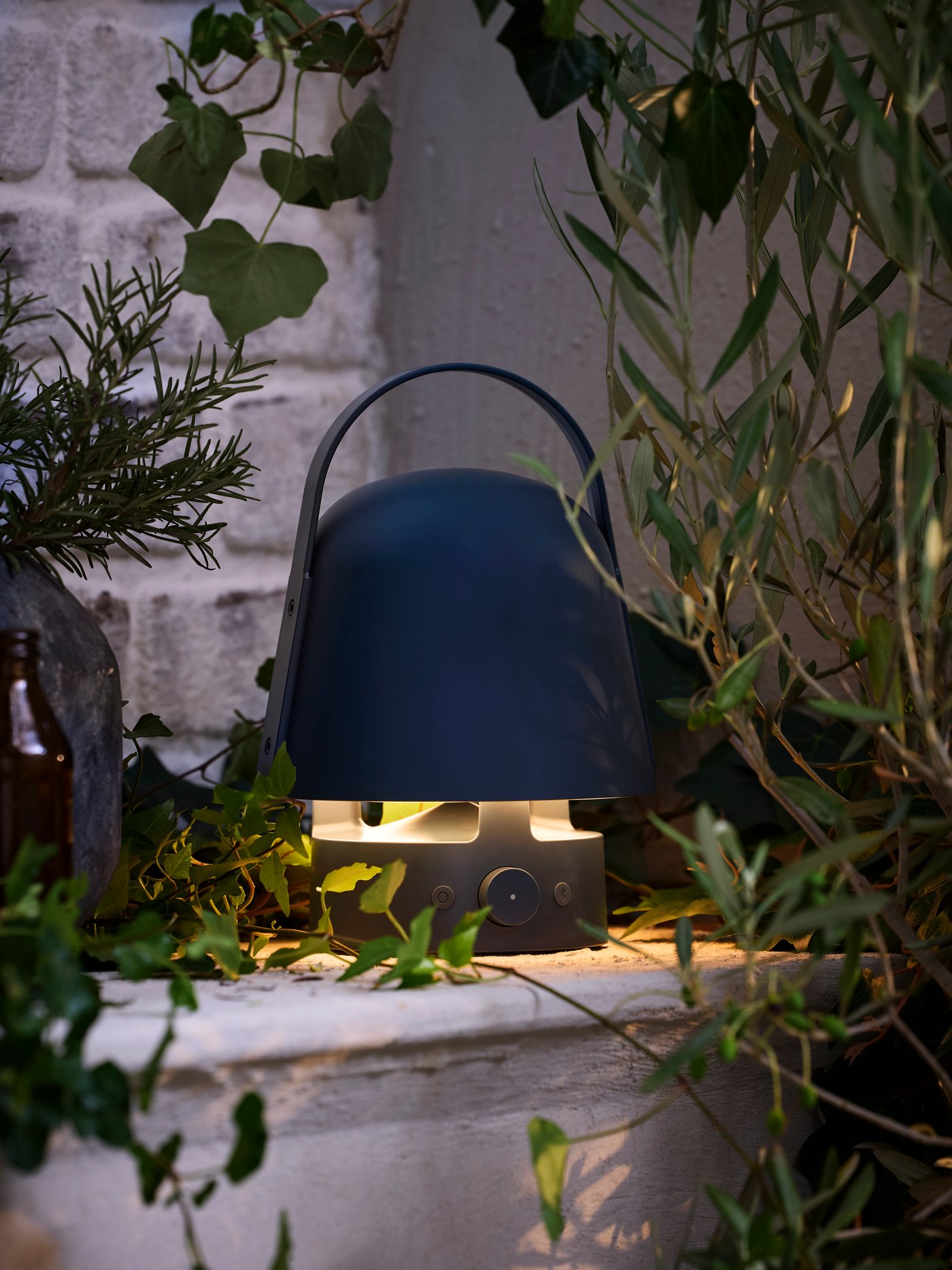 منظر للفناء عند الغروب به مصباح بسماعة بلوتوث خارجي VAPPEBY أزرق موضوع في زاوية. ينشر ضوءاً ناعماً ودافئاً.