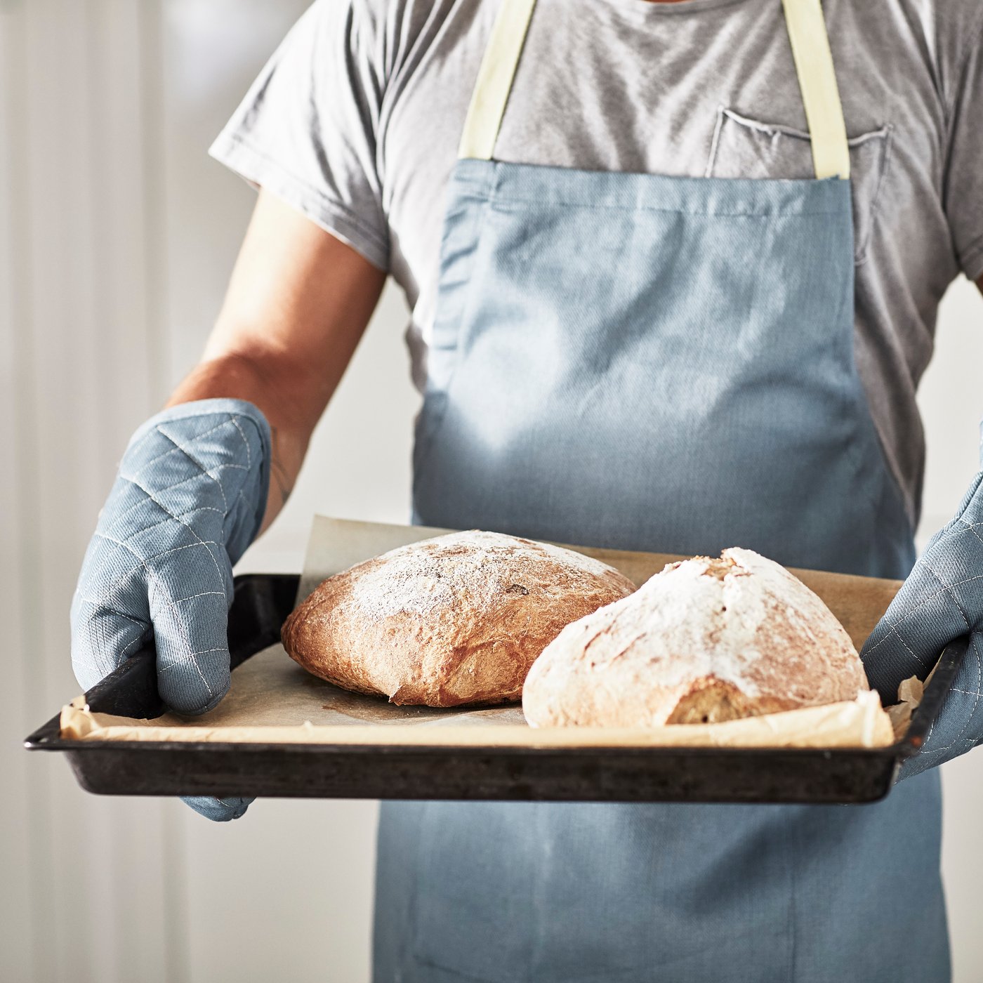 En person med förkläde och grytvantar håller i en plåt med nybakat bröd.