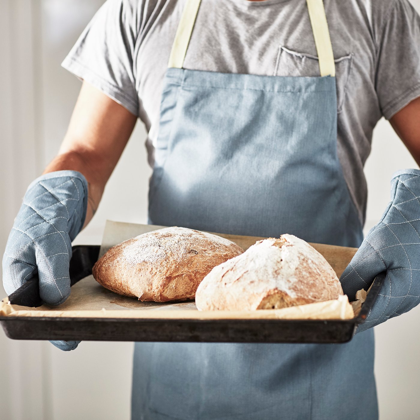 En person iført et lyseblåt forklæde og SANDVIVA ovnhandsker holder en bageplade med bagepapir og to friskbagte brød.