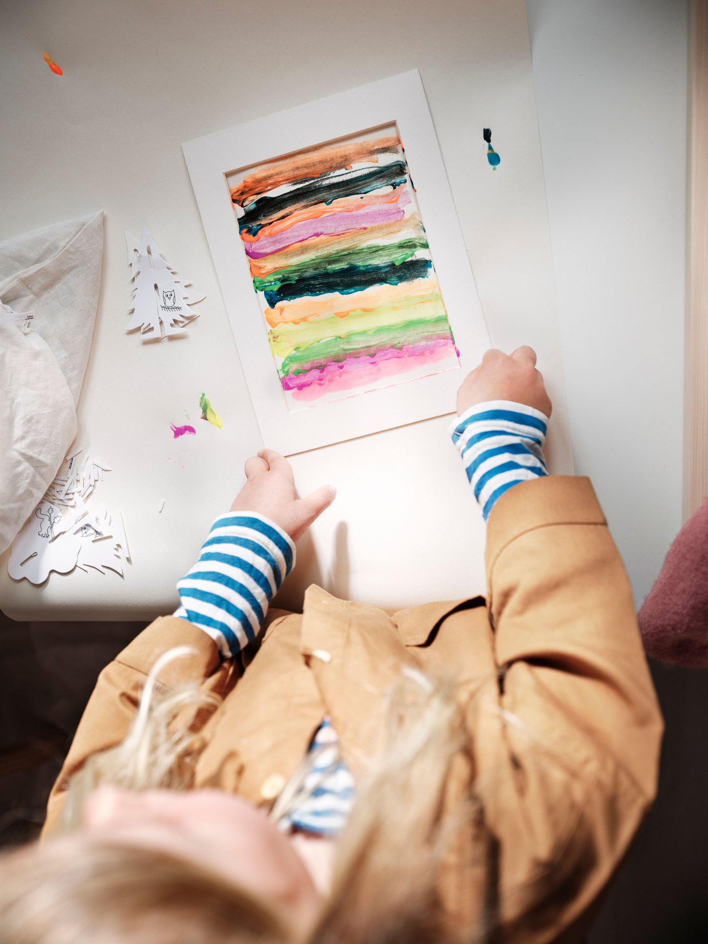 Laps, kellel on beež jakk ja sinivalgetriibulised varrukad, istub laua taga ning hoiab käes värviküllast, mitmekihilist akvarellimaaliga paberilehte.