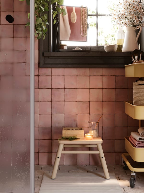 A pink bathroom has an OPPEJEN shower door to the left, a windowsill with a mirror on it, a step stool and a RÅSKOG trolley.