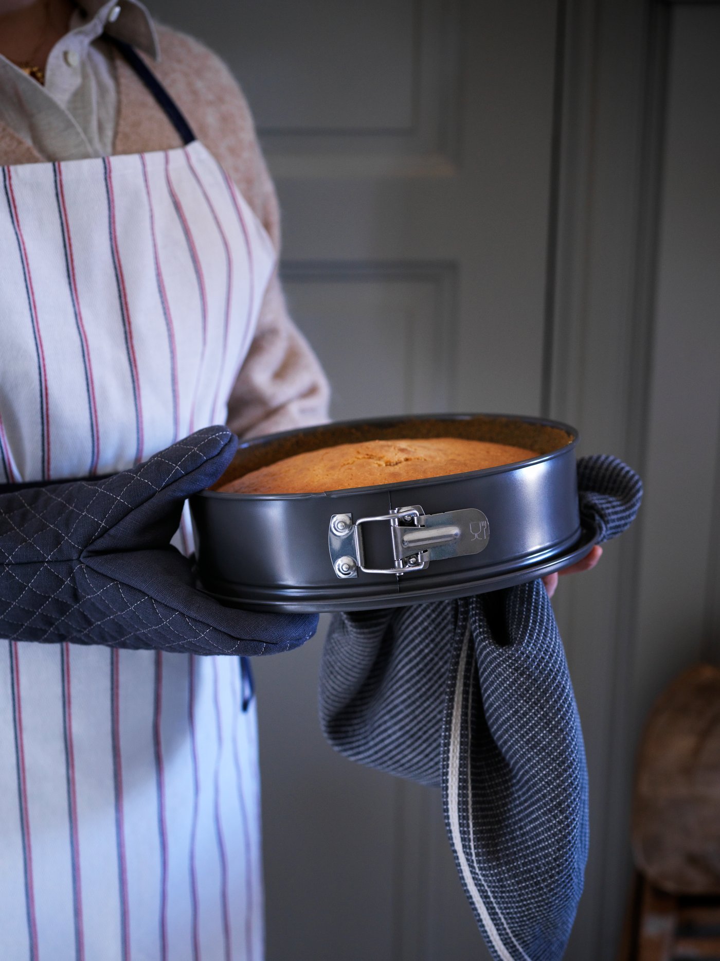 A person wearing a HILDEGUN apron and MARIATHERES oven glove holds a baked cake in the grey HEMMABAK springform pan.