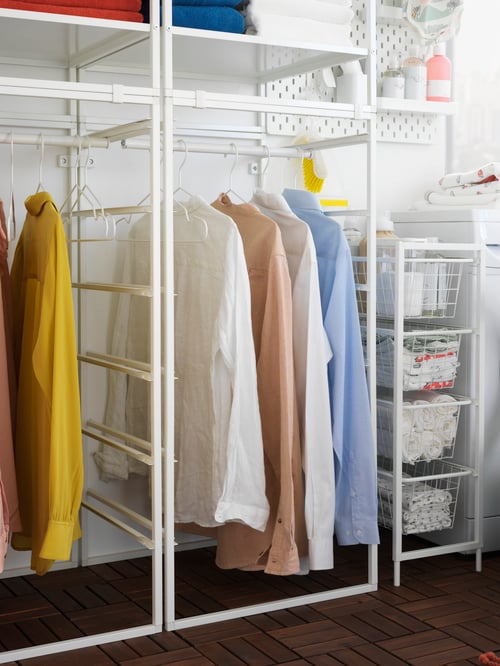 Colourful shirts hang at the bottom of a white JONAXEL white shelving unit with clothes rails, by baskets in a laundry area.