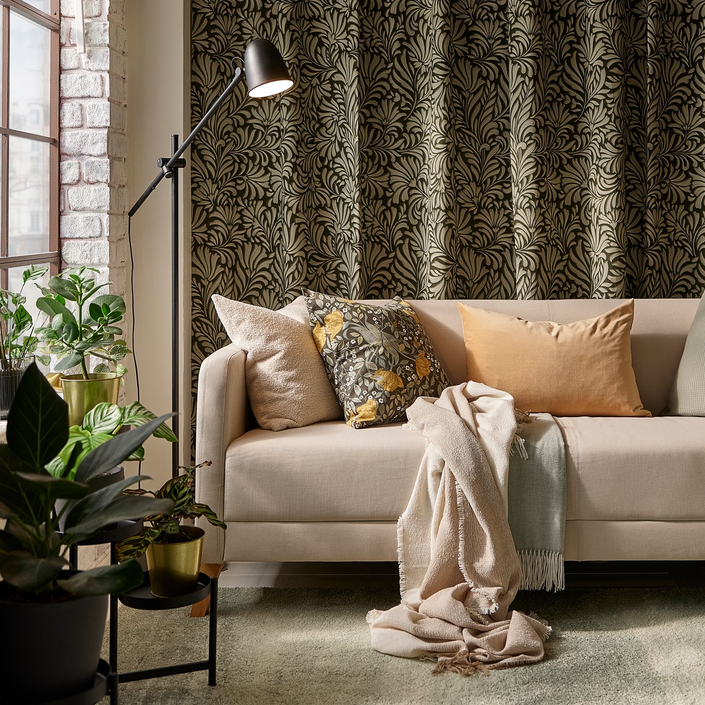 Divano LINANÄS beige con vari cuscini, su un tappeto grigio-verde chiaro; sullo sfondo, tende con motivo a foglie a ricoprire tutta la parete.
