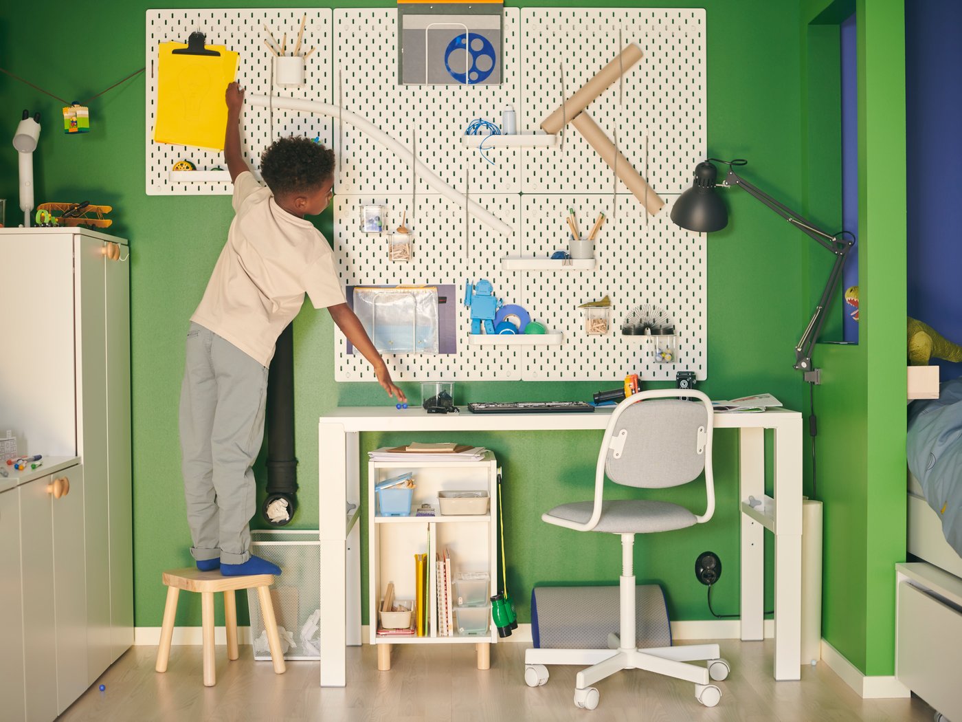 Ein Kind steht auf einem Hocker vor einer SKÅDIS Lochplatte, die über einem weißen PÅHL Schreibtisch an einer grünen Wand hängt.