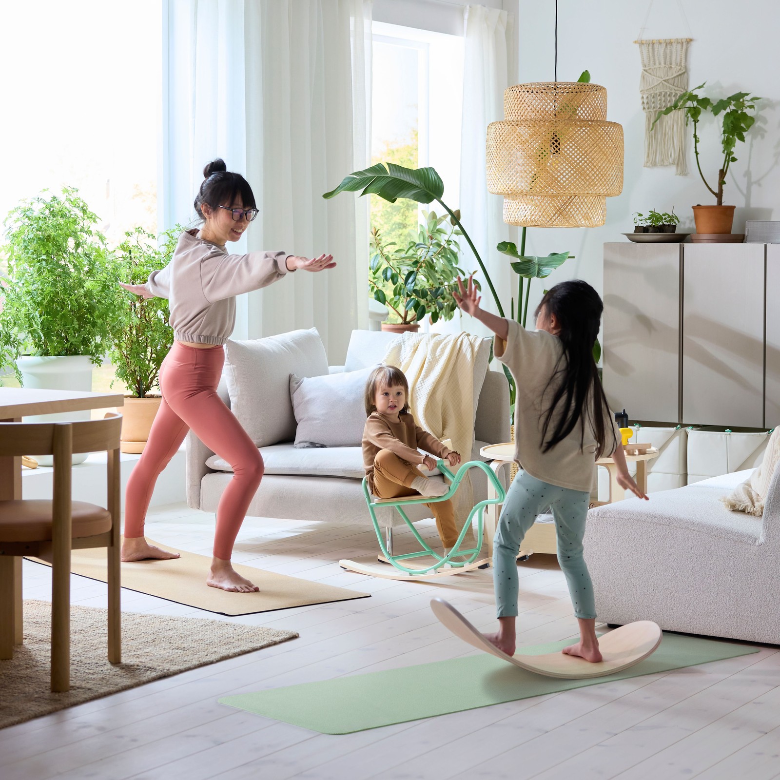 In a living room, a parent practices yoga as their children play on a GREJSIMOJS balance board and GREJSIMOJS rocking-bird.