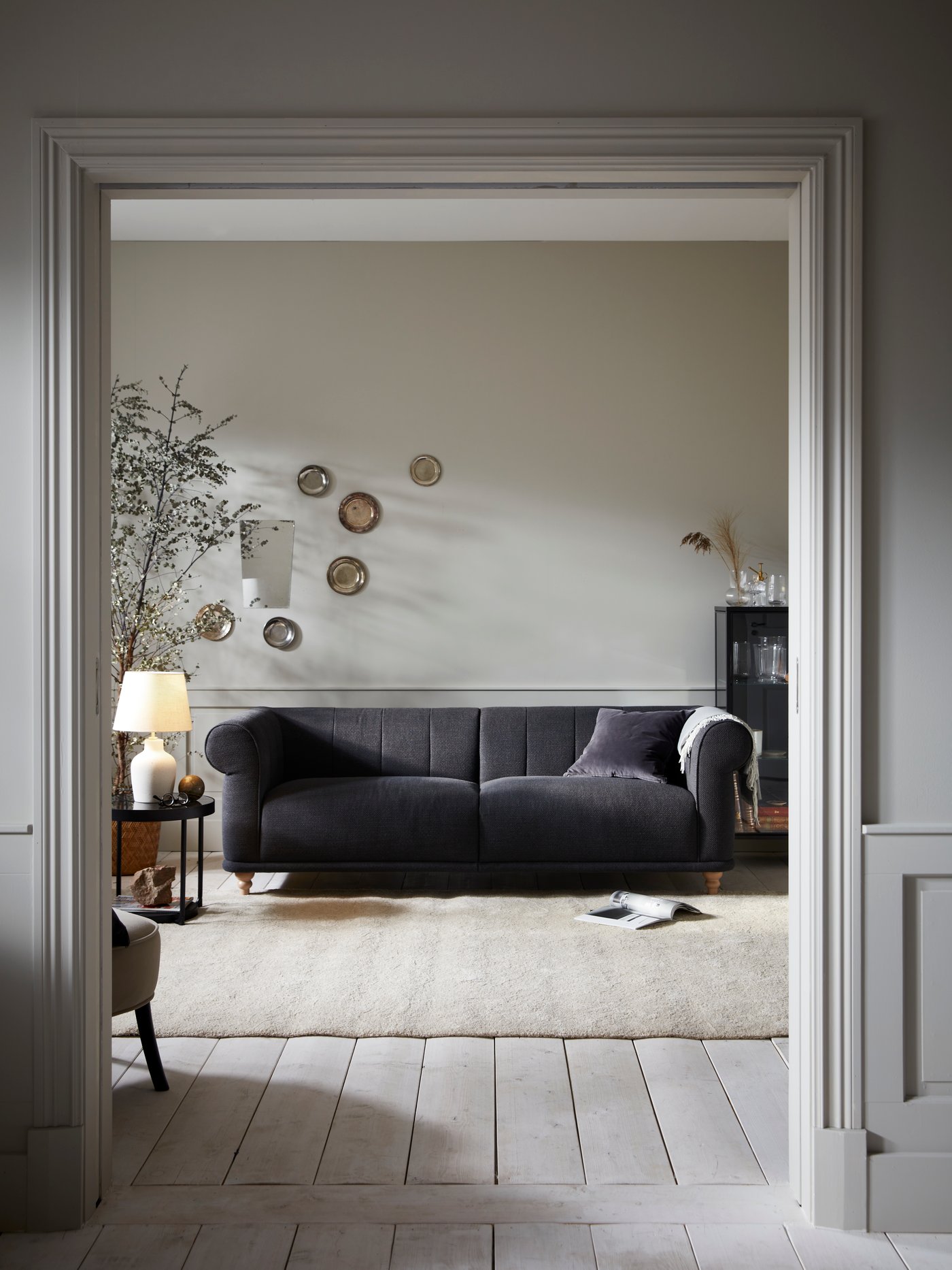 A VISKAFORS 3-seater sofa stands in a living room. A HOLMVI plaid is draped over an armrest. A switched-on table lamp can be seen on a side table next to the sofa.