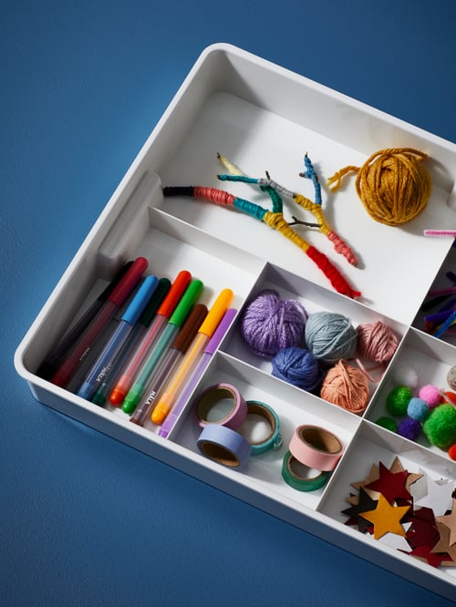 A white KUGGIS insert with eight compartments filled with MÅLA mixed-colours felt-tip pens, string and crafts accessories.