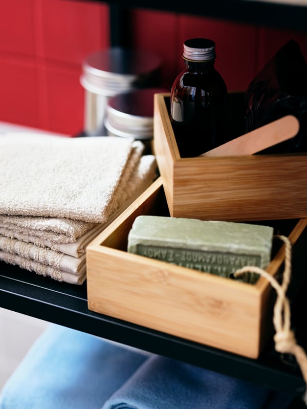 Folded towels, jars and bottles, and bathroom accessories in a pair of TAVELÅN trays share a shelf of an ENHET cabinet.
