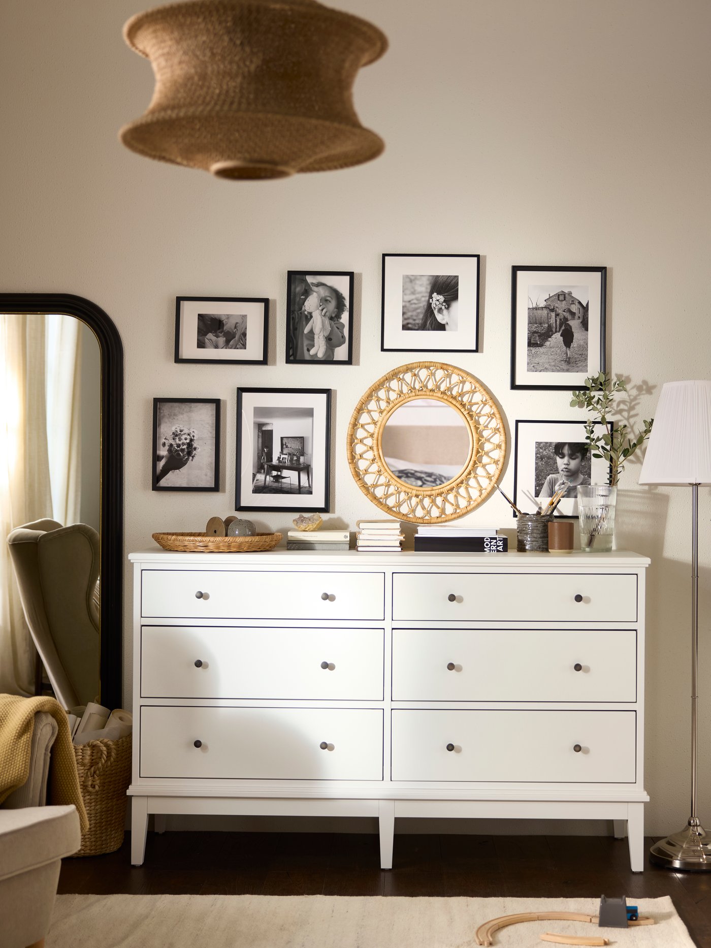 Bílá komoda IDANÄS se šesti zásuvkami, na které jsou umístěné různé dekorativní předměty, a spousta zarámovaných fotografií a zrcadlo na stěně nad touto komodou.