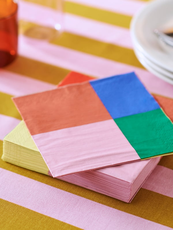 A stack of colourful TESAMMANS paper napkins is on a table with TESAMMANS pre-cut fabric used as a tablecloth.