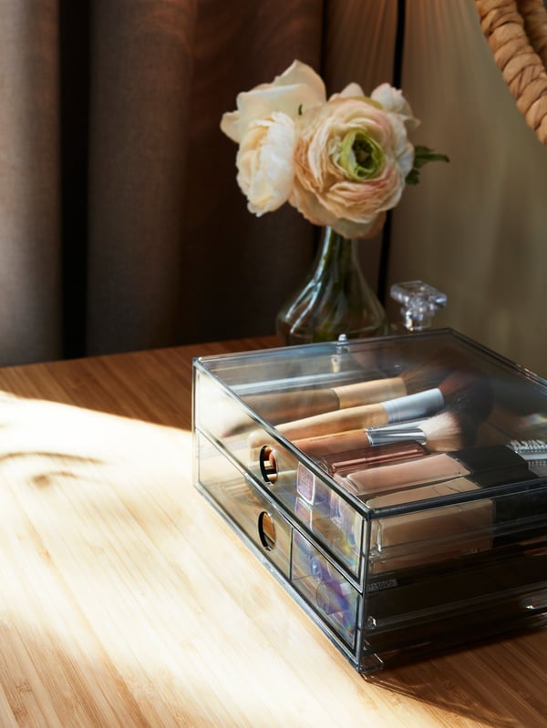 Des articles de maquillage sont rangés dans une minicommode à deux tiroirs GODMORGON fumés à côté d'un vase à fleurs et d'une coiffeuse.