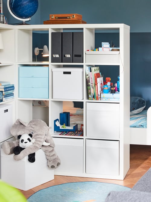 Deux étagères blanches KALLAX contenant des boîtes et des jouets sont utilisées comme séparateur de pièce. Derrière, se trouve un lit pour enfant.