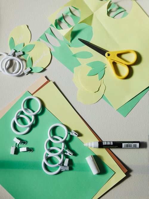 Bucăți de hârtie de culoare verde și galbenă așezate pe o masă, împreună cu foarfece și inele pentru draperii cu clips.