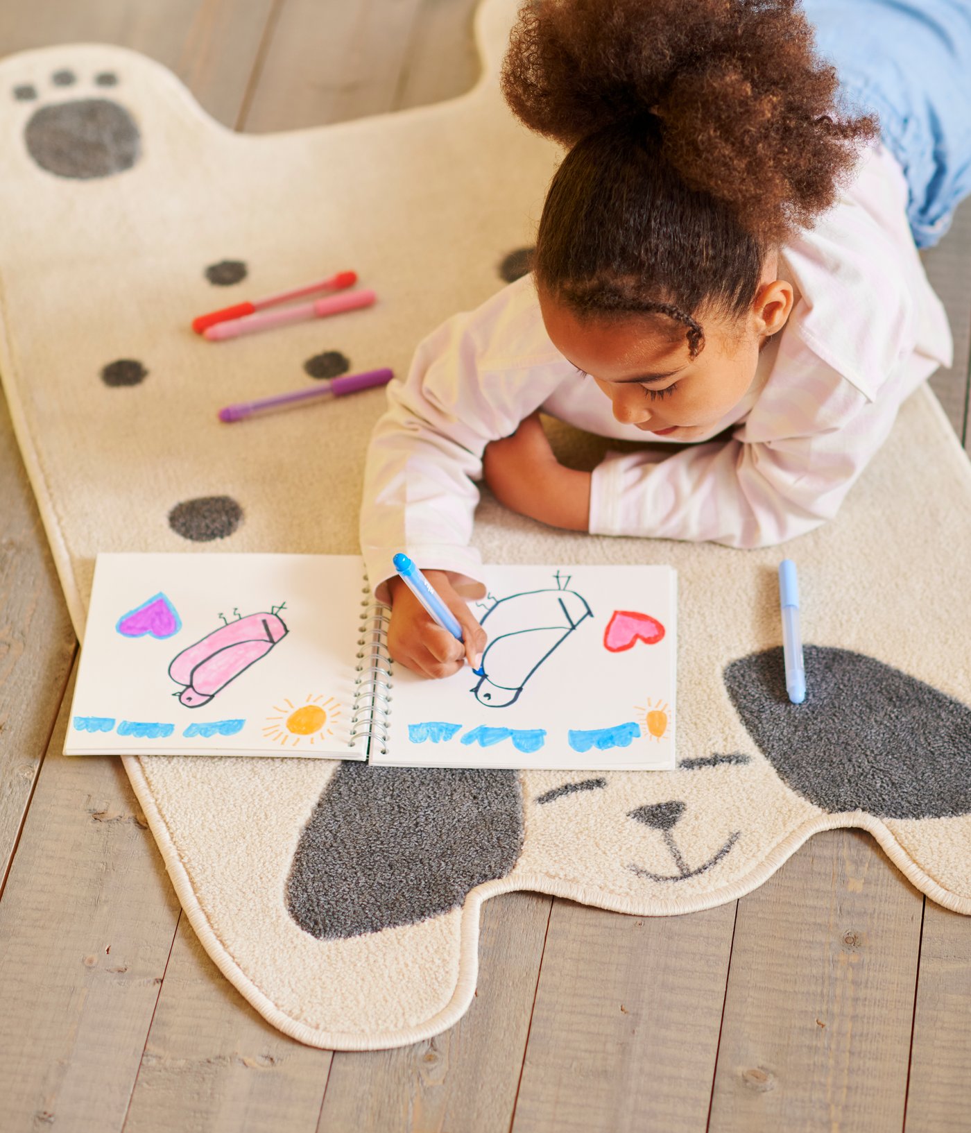 A child is drawing in a notebook using colourful pens and smiling, lying on a DRÖMSLOTT dog-shaped rug.