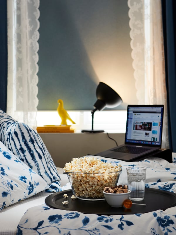 Un letto con cuscini e biancheria bianca e azzurra. Sopra il letto, c'è un vassoio con pop corn, salatini e bevande. A lato, un laptop aperto.