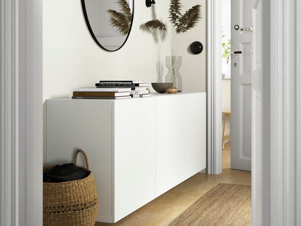 Minimalistisk hall med vit BESTÅ skänk, spegel, böcker, vas med torkade växter och korg på trägolv med matta.
