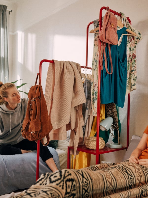 A red clothes rack is used to divide a room, with a bed on either side. Two young adults are sitting on the beds.