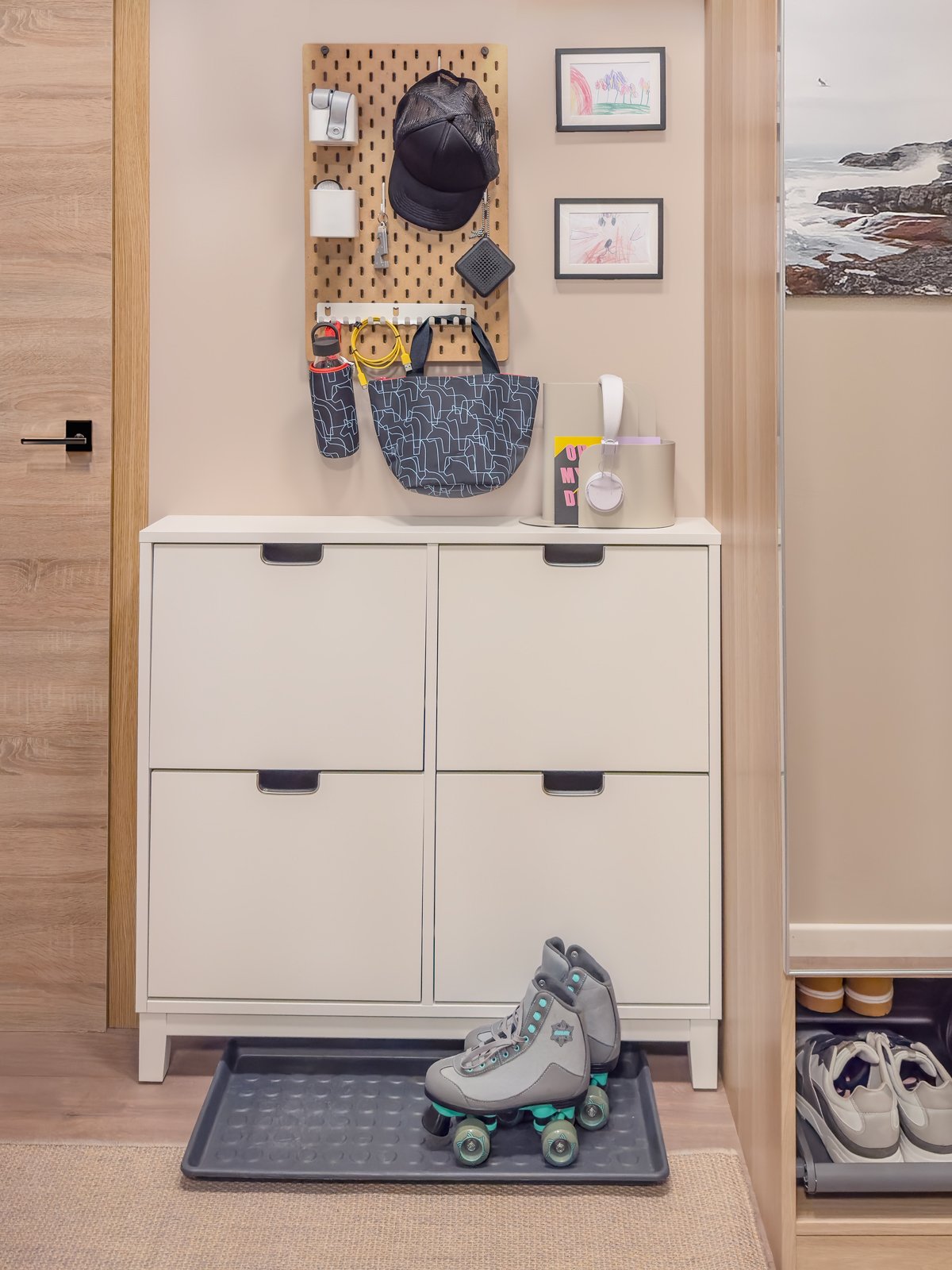 Vit hallbyrå med fyra lådor, ovanför en väggpanel med krokar och små hyllor. På golvet står ett par rullskridskor på en gummimatta.