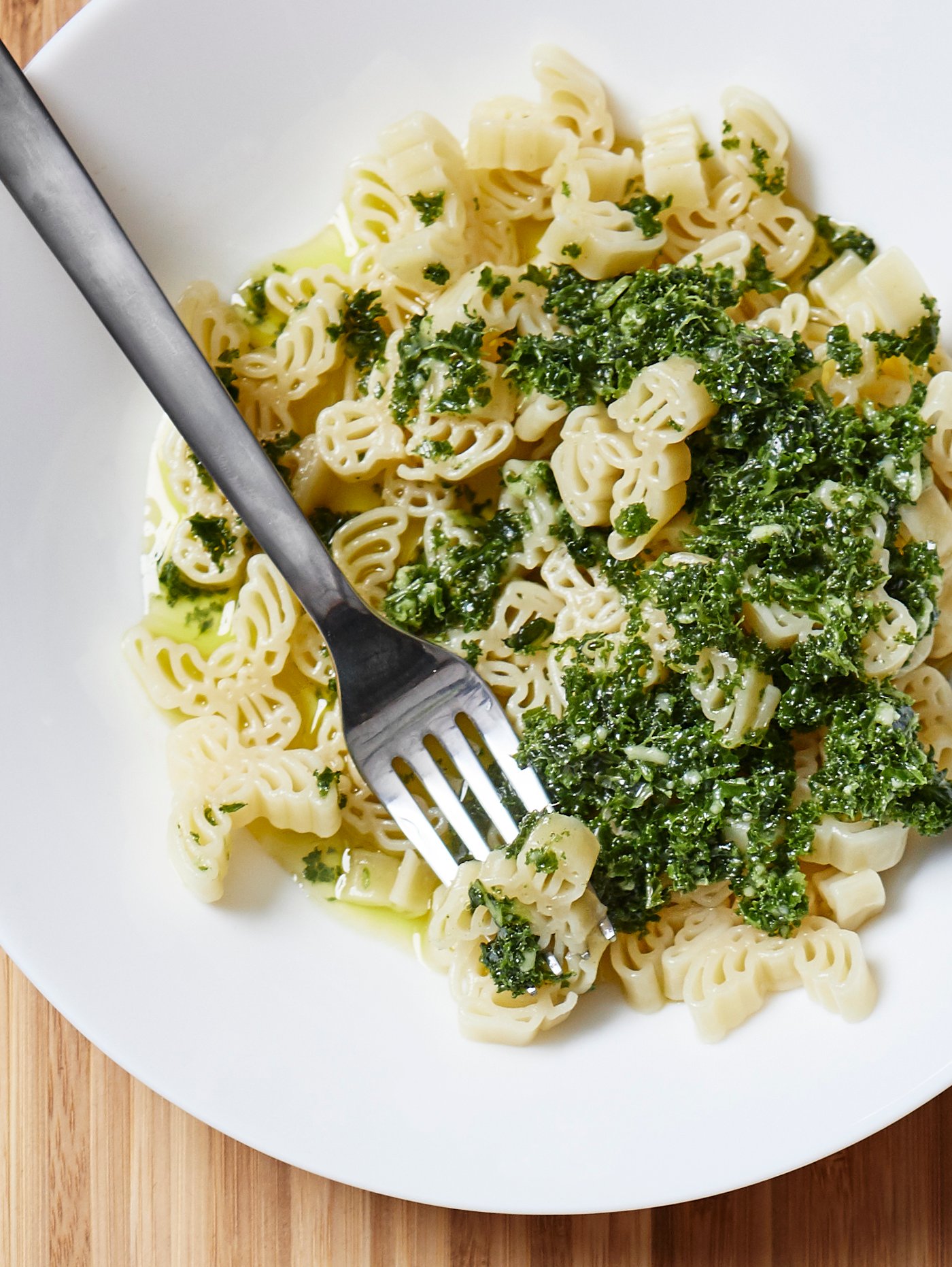 Fotografía cenital de pasta y guarnición verde en un plato hondo blanco. Hay un tenedor de metal colocado en el plato hondo.