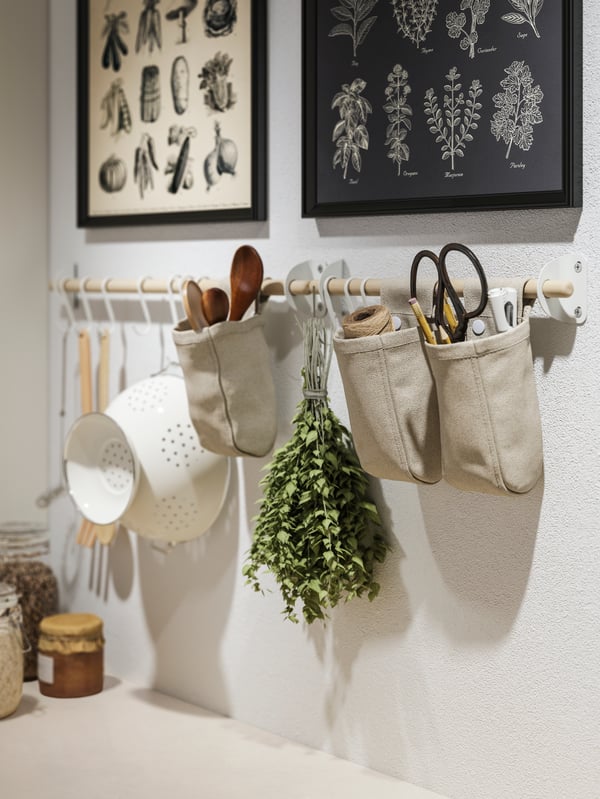 Küchenwand mit zwei gerahmten botanischen Illustrationen von Gemüse und Kräutern. Darunter eine Holzstange mit hängenden Küchenutensilien, darunter ein weißes Sieb, Holzlöffel, eine Schere und ein Bündel frischer Kräuter. Links unten stehen Vorratsgläser.