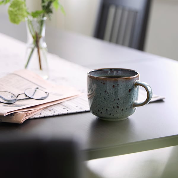 A beige-green GLADELIG mug rests on a table next to a newspaper and glasses, with a vase of greenery in the background.