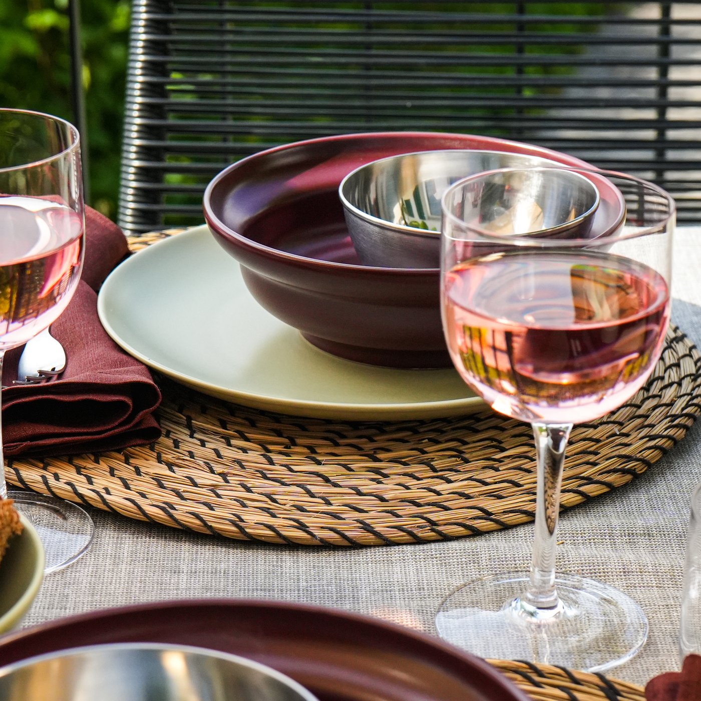 Une table dressée avec des verres à vin rosé, de la vaisselle foncée posée sur des sets de table en raphia et des tranches de pain noir, idéale pour un repas convivial.