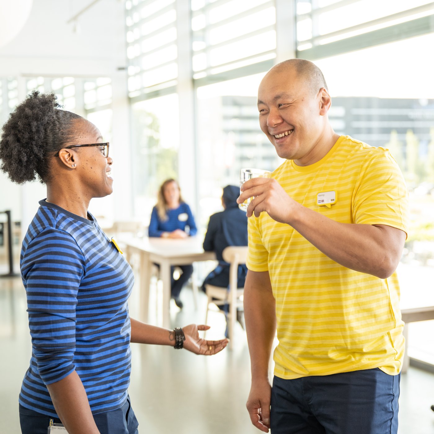 Un homme et une femme en T-shirts Ikea rayés conversent dans un espace lumineux et moderne avec de grandes fenêtres ; À l’arrière-plan, des gens à des tables, certains en uniforme.