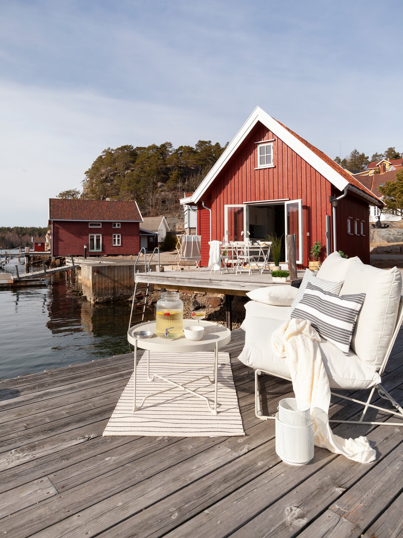 En rød norsk hytte beliggende ned til en søbred med en indbydende indrettet træterrasse med havemøbler og tekstiler.