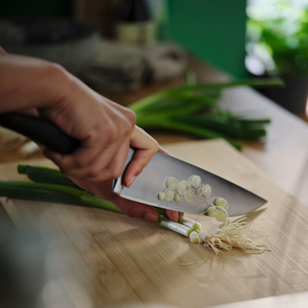 Two hands are holding and cutting vegetables on a bamboo APTITLIG chopping board with a black VÖRDA cook’s knife.