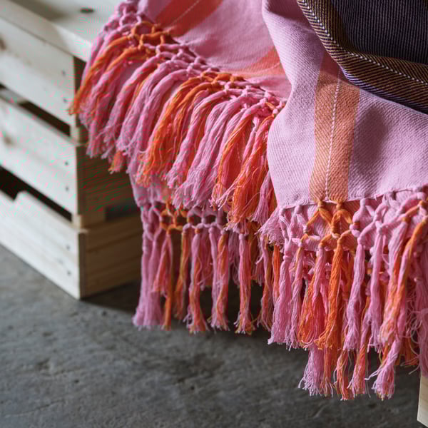A MÄVINN throw with stripes in pink and orange and handtied tassels is displayed on a wooden box.