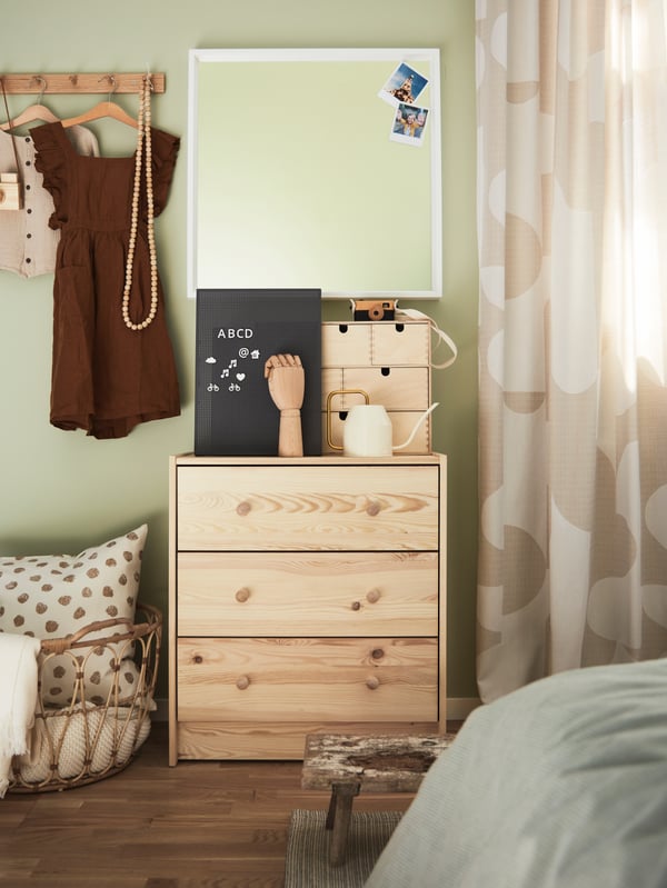A pine RAST chest of drawers with various items on top and a white mirror above, beside a basket with a cushion.