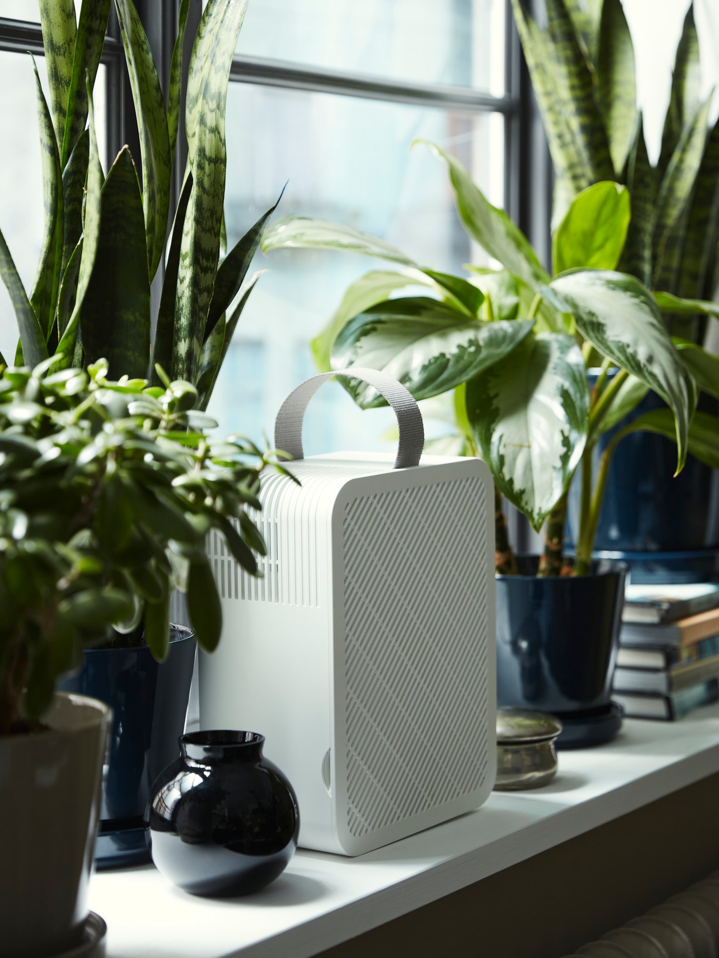 Ein UPPÅTVIND-Luftreiniger steht auf einer Fensterbank neben einigen Pflanzen in Pflanztöpfen, Büchern und anderen Gegenständen.