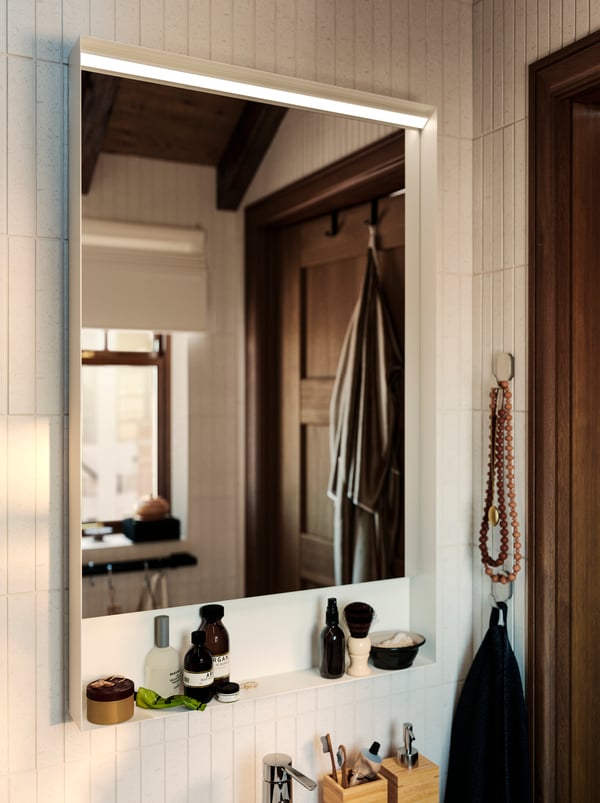 A FAXÄLVEN mirror with built-in lighting has bathroom products placed on a shelf and there are two hooks mounted on the wall.