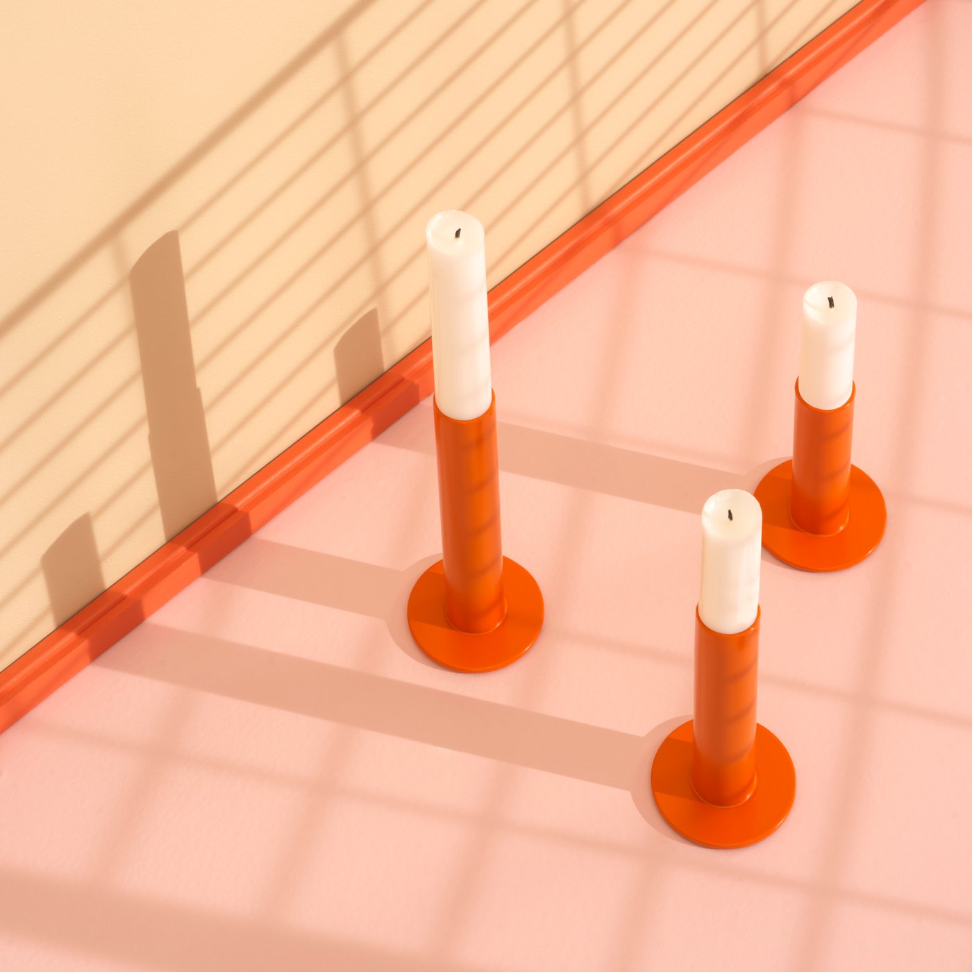 Set tiga bekas lilin keluli TUVKORNELL berwarna jingga dengan lilin berwarna putih terletak di atas lantai berwarna merah jambu pucat di dalam studio.