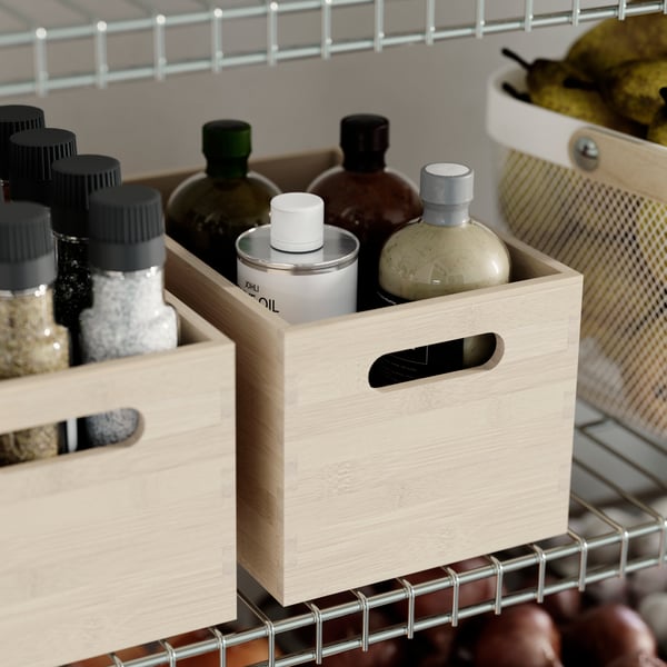 Una caja de almacenamiento UPPDATERA de madera clara, colocada sobre una estantería OMAR y llena de botellas junto a tarros y otros artículos de despensa.