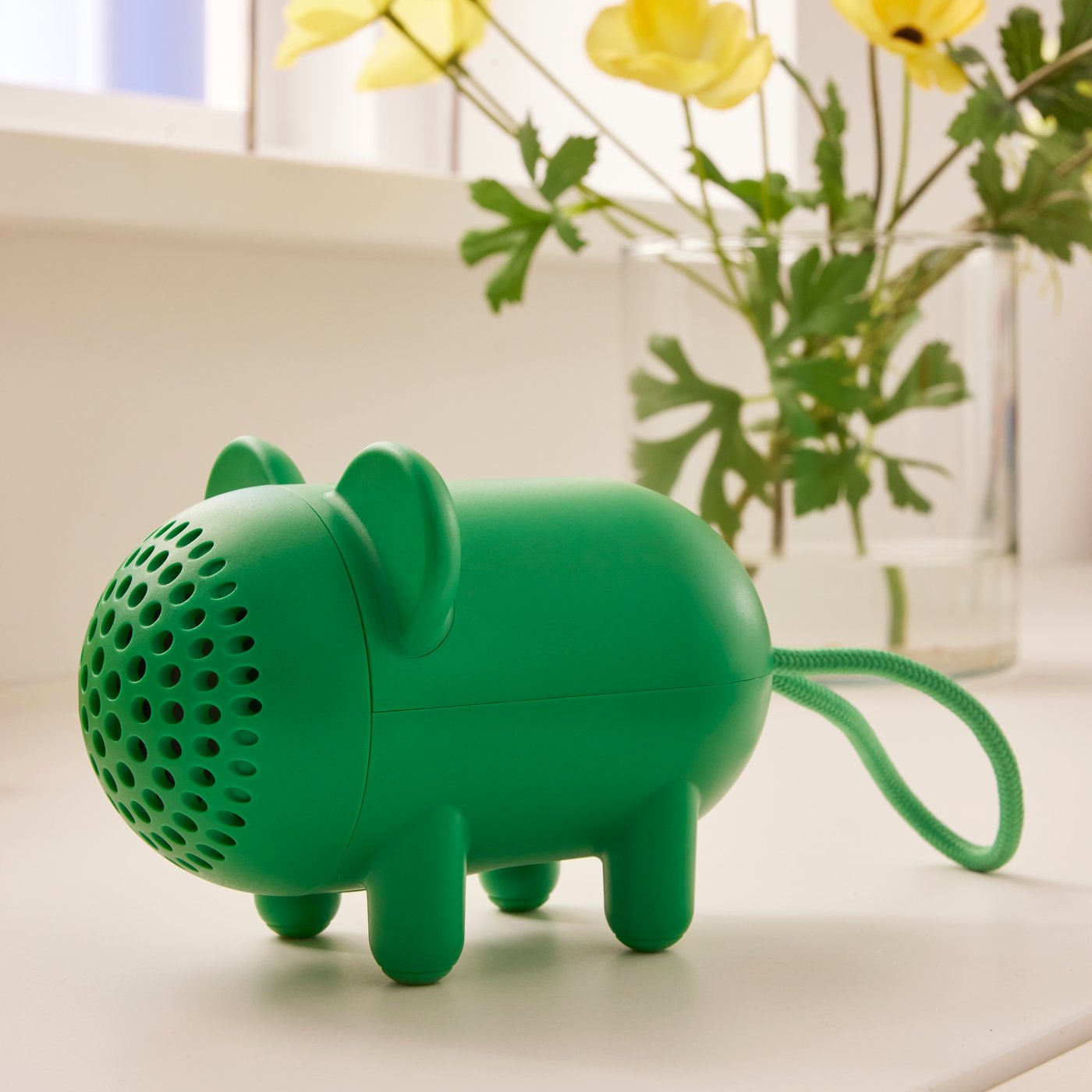 Un altoparlante Bluetooth verde a forma di maialino con la parte anteriore traforata che ricorda un muso. È appoggiato su una superficie chiara accanto a un vaso con fiori gialli.