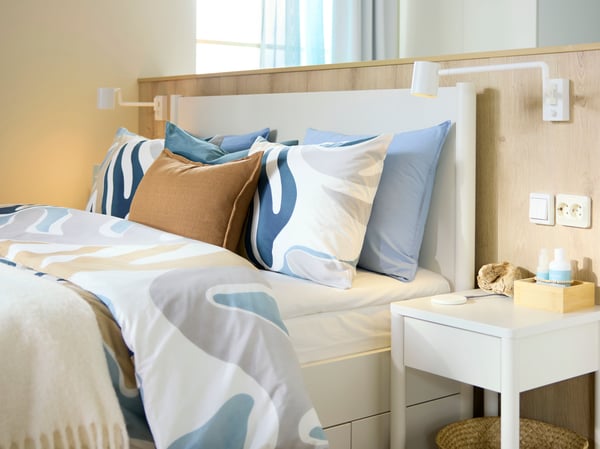 A bed has a leafy-patterned MYGGLASVINGE quilt cover and pillowcases on it, against a low wall in a light-filled hotel room.
