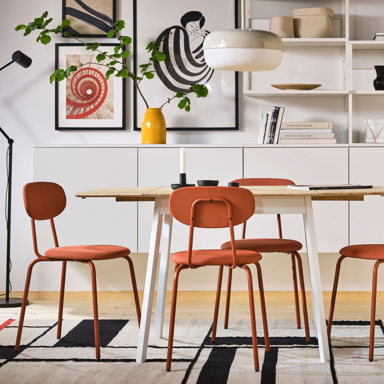 Svetao trpezarijski kutak s uramljenim umetničkim delom, PLATSA spojenim policama i crveno-smeđim ÖSTANÖ stolicama i IKEA PS stolom na rasklapanje.