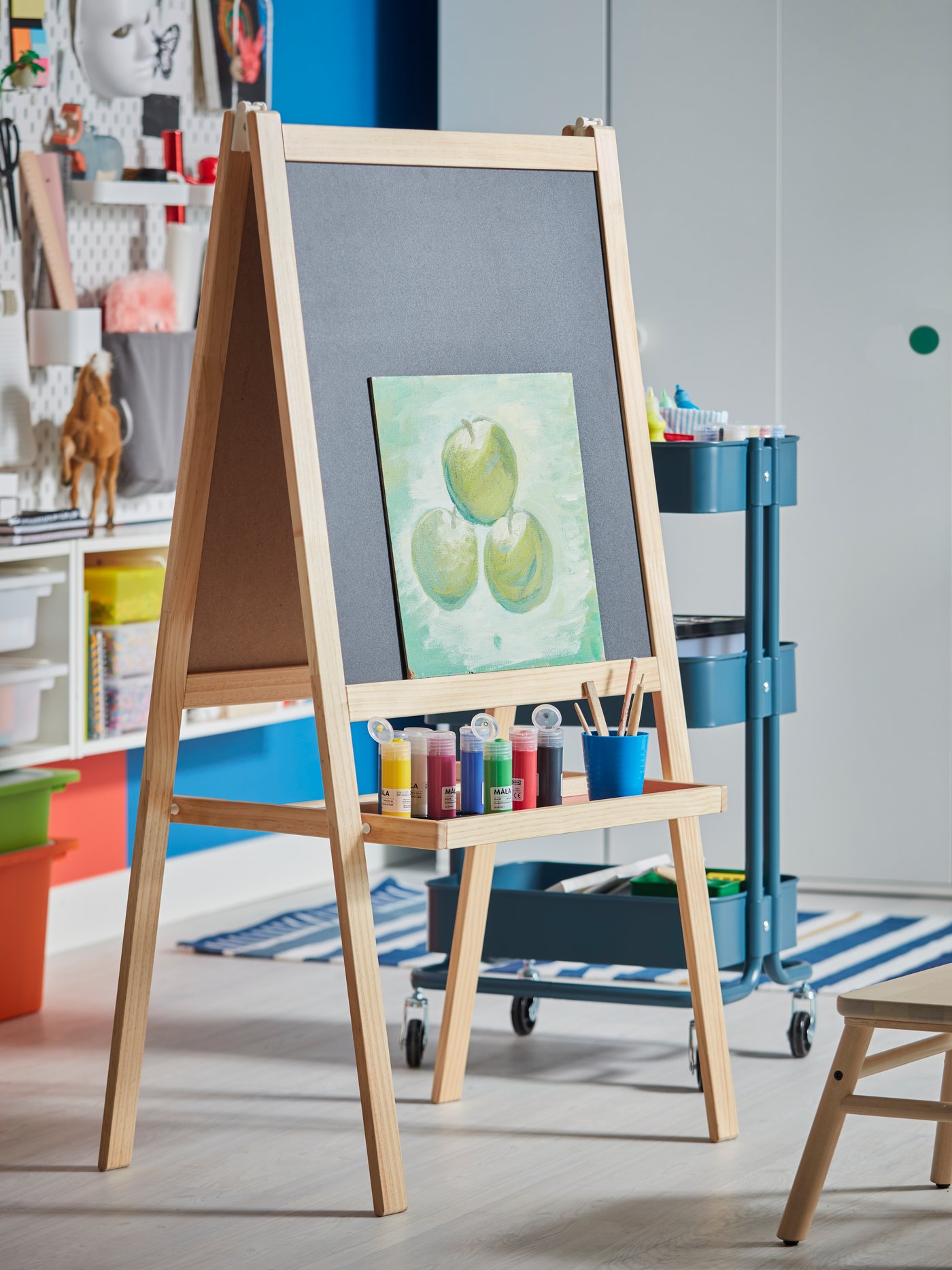A MÅLA easel with a painting of apples and colorful MÅLA paints and paintbrushes stands in a child’s bedroom.