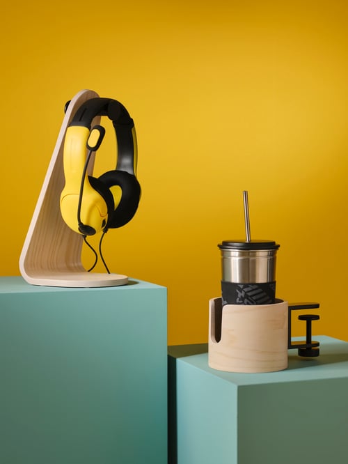 A LÅNESPELARE headset stand with a pair on headphones on top, next to a mug holder with a mug with lid and straw on top.