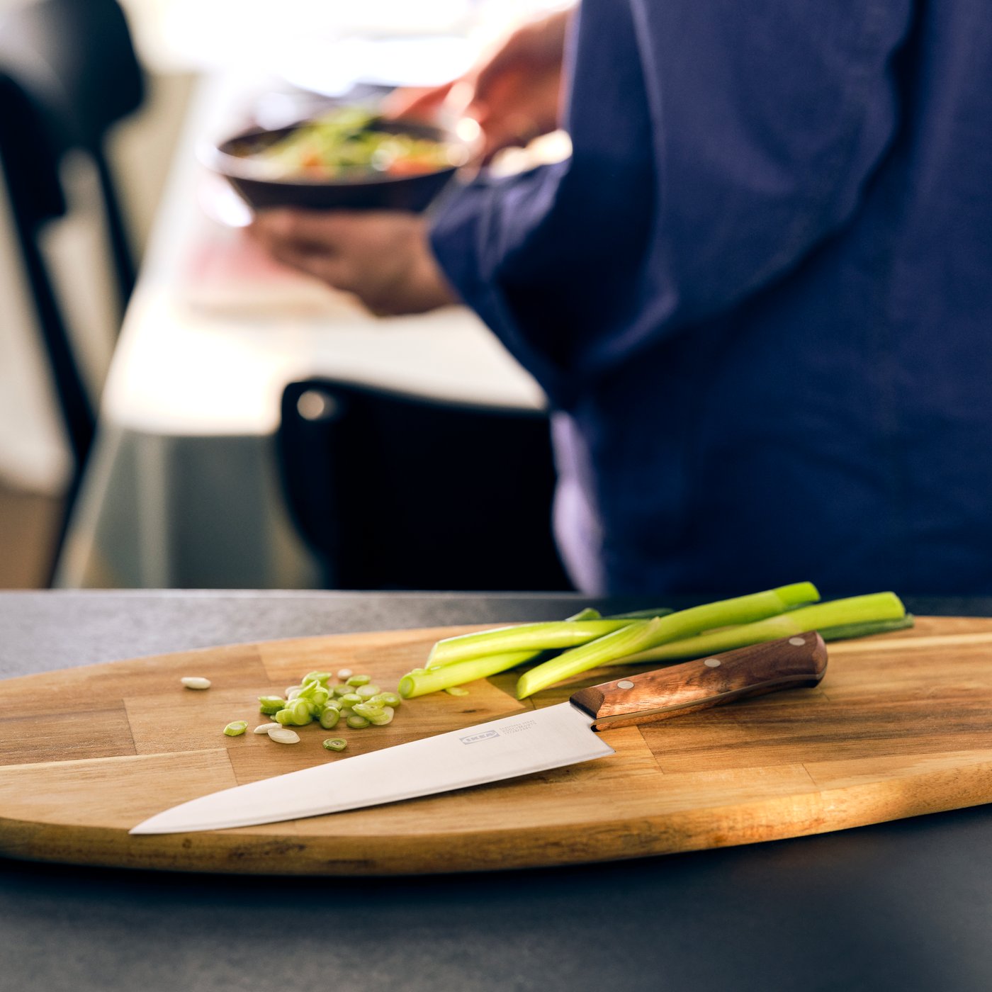 Нож VARDAGEN с деревянной ручкой лежит на овальной разделочной доске рядом с зеленым луком.