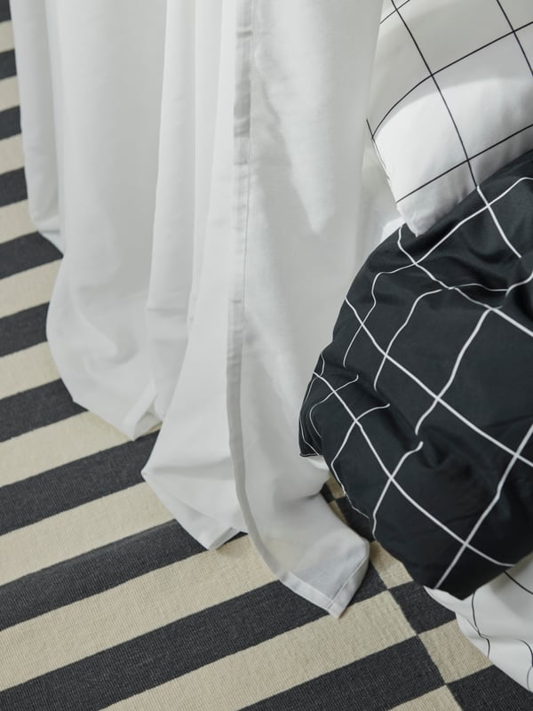 The corner of a duvet and pillow covered in VITKLÖVER bed linen beside the bottom of a sheer white ROSENROBINIA curtain.